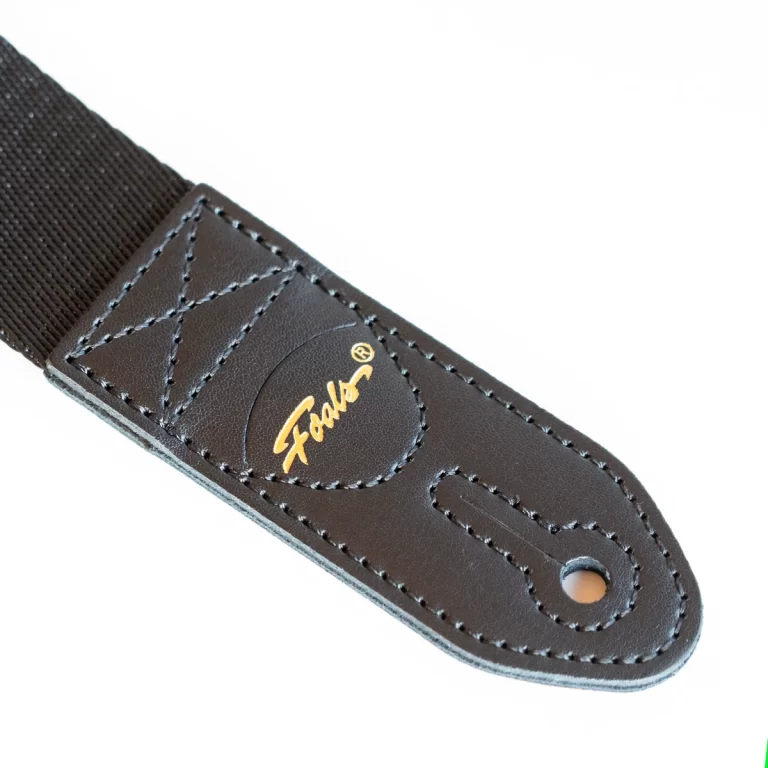 Close-up of a brown leather guitar strap end with stitched edges and a small gold logo patch beside a hole for the strap button. ขายราคาพิเศษ