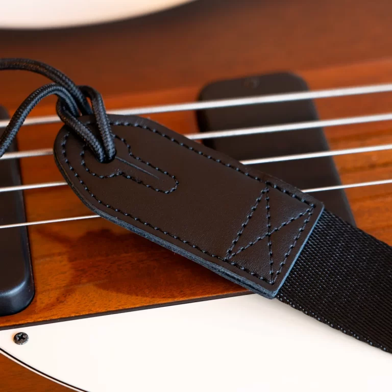 Close-up of a black leather guitar strap with stitching attached to the body of an electric guitar, showing strings and a white pickguard edge nearby ขายราคาพิเศษ