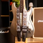 Metallic electric guitar with a white pickguard, draped by a decorative floral strap in a music setup. ขายราคาพิเศษ