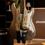 Golden electric guitar on a stand with a colorful patterned strap and a white pickguard, in a music room setting with an amp in the background ขายราคาพิเศษ