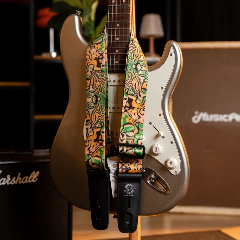 Golden electric guitar on a stand with a colorful patterned strap and a white pickguard, in a music room setting with an amp in the background ขายราคาพิเศษ