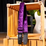 Purple velvet guitar strap with black leather ends, hanging over a wooden crate with a white electric guitar visible in the background. ขายราคาพิเศษ