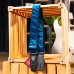 Blue denim guitar strap with leather ends hanging on a wooden crate, with a blurred white guitar in the background. ขายราคาพิเศษ