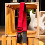 Red guitar strap with black leather ends draped over a wooden crate, with a white electric guitar visible in the background hastily blurred in a store setting. ขายราคาพิเศษ