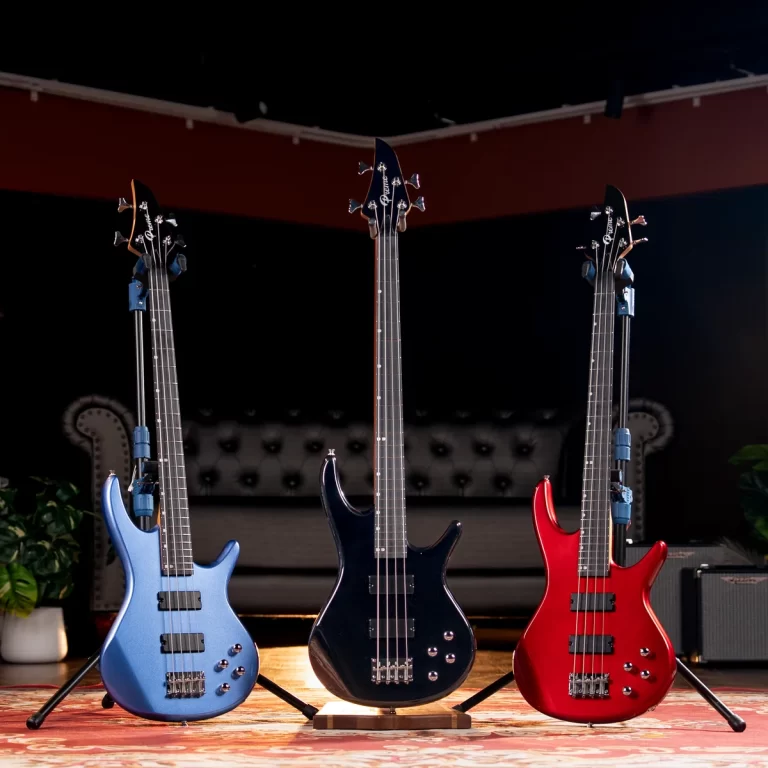 Three electric bass guitars in blue, black, and red stand on a stage with a patterned rug and dark backdrop. ขายราคาพิเศษ