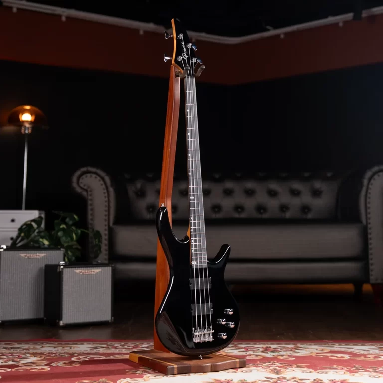Black electric bass guitar on a wooden stand in a dim living-room studio with a tufted sofa and small amplifiers in the background. ขายราคาพิเศษ