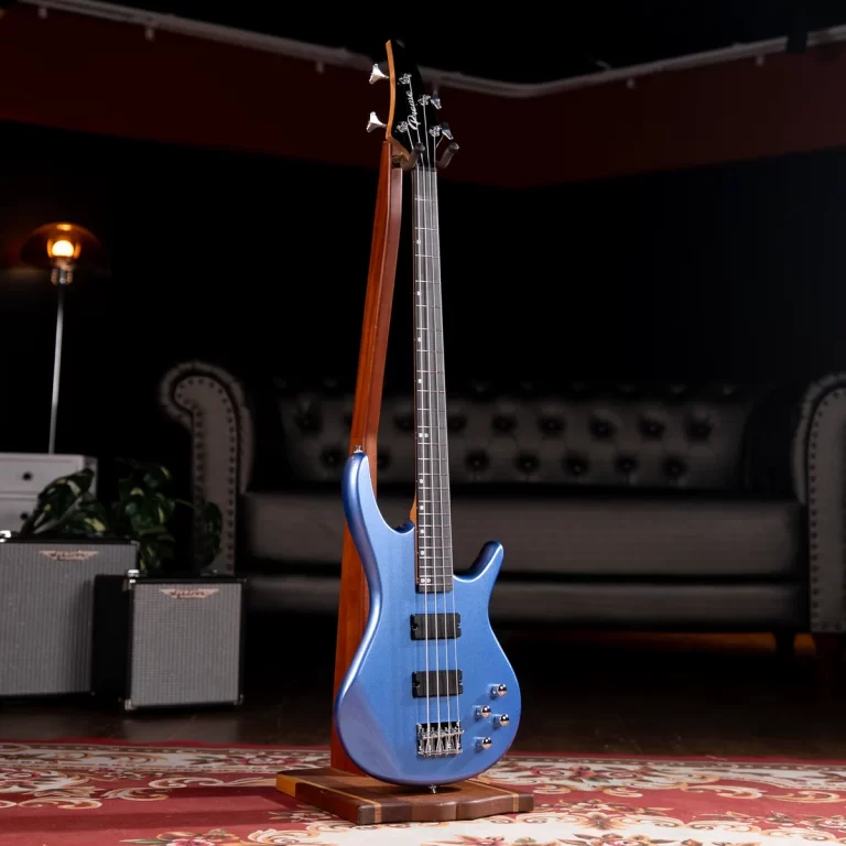 Blue electric bass guitar on a wooden stand, standing on a patterned rug in a home studio with amps and a tufted sofa in the background. ขายราคาพิเศษ