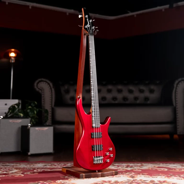 Red electric bass guitar on a wooden stand in a dimly lit living-room studio with a patterned rug behind it. ขายราคาพิเศษ