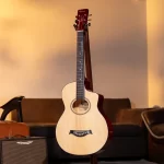 An acoustic guitar on a stand in a warmly lit living room, with a small amplifier and couch visible in the background. ขายราคาพิเศษ