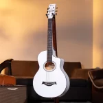 White acoustic guitar on a stand in a cozy living room with warm lighting and a brown sofa in the background. ขายราคาพิเศษ