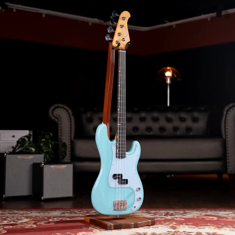 Light blue electric bass guitar on a stand in a cozy studio/living room with a tufted sofa and warm lamp in the background. ขายราคาพิเศษ