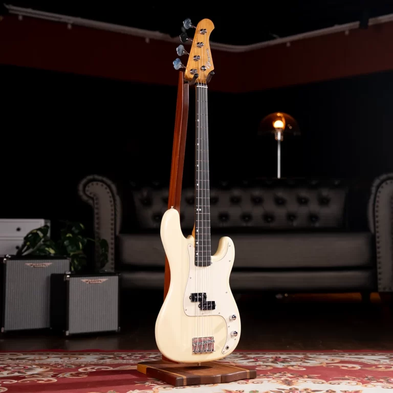 Cream-colored electric bass guitar on a wooden stand in a dim living-room setting with a tufted sofa and small amplifiers in the background. ขายราคาพิเศษ