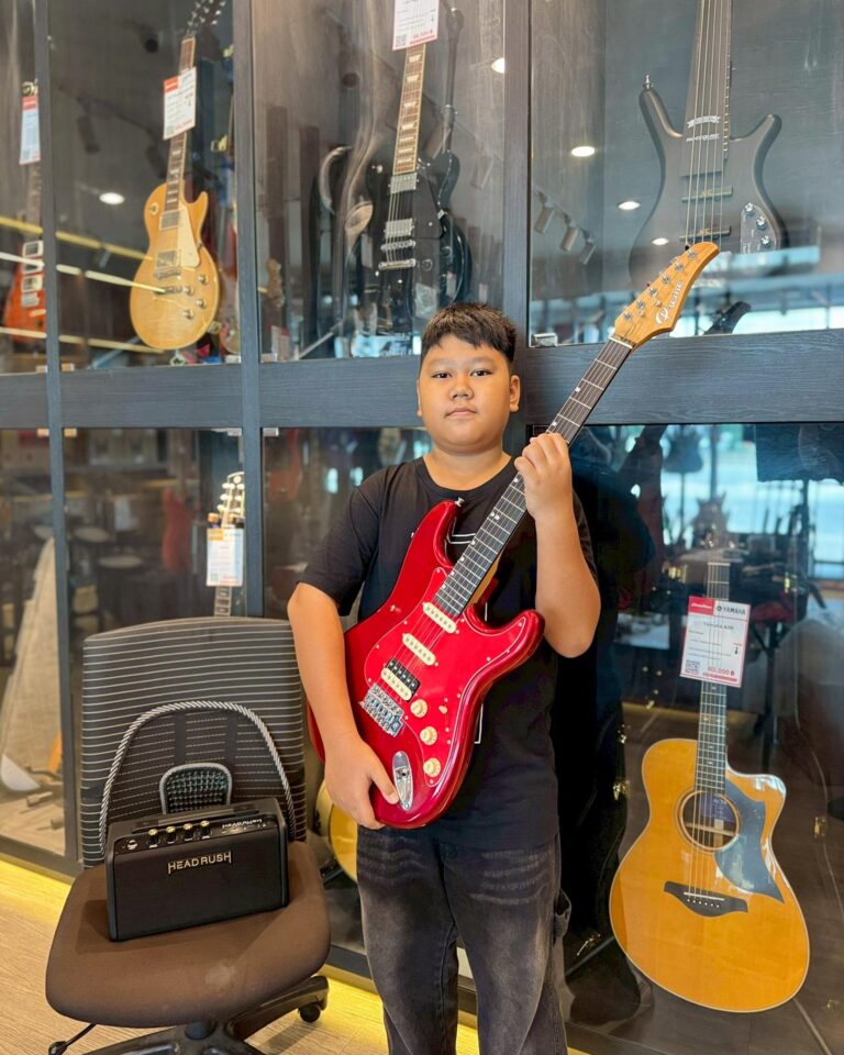 Boy in a music store posing with a red electric guitar in front of a glass display of guitars behind him.