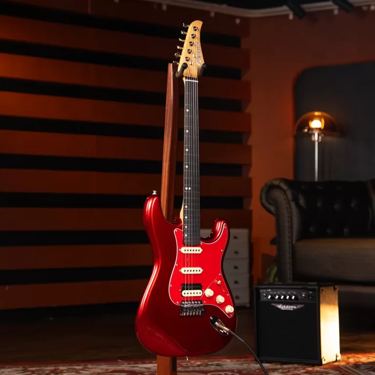 Red Stratocaster-style electric guitar on a stand in a recording studio, plugged into a small amplifier, with orange acoustic panels and a sofa in the background. ขายราคาพิเศษ