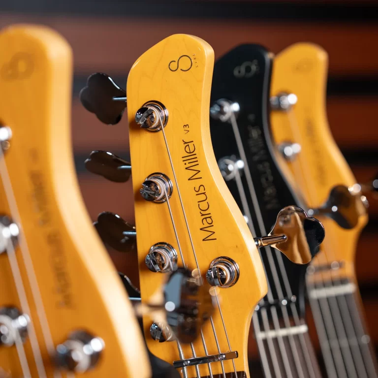 Close-up of yellow Marcus Miller V3 guitar headstock with tuning pegs in focus. Necks sit in a row, warm-toned lighting. ขายราคาพิเศษ