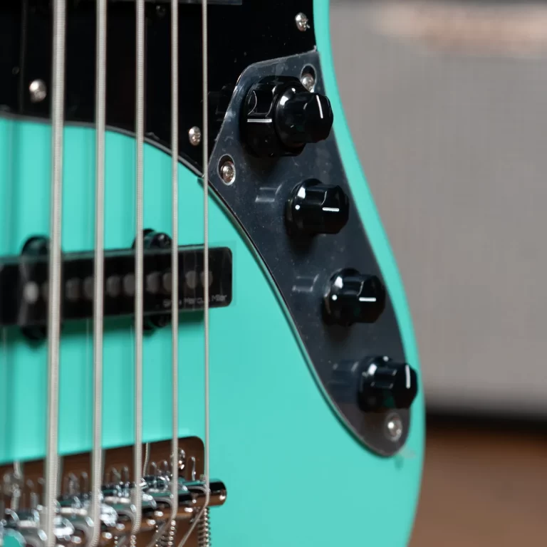 Close-up of a turquoise electric bass guitar body, showing the black control plate, multiple knobs, strings, and chrome bridge components up close. ขายราคาพิเศษ