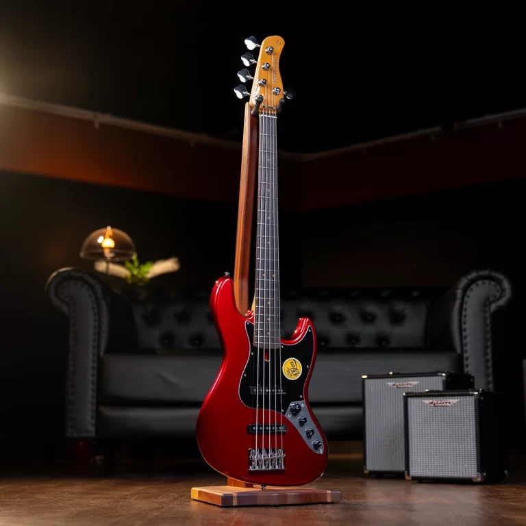 Red electric bass guitar on a stand in a dim studio, with a tufted sofa and two amplifiers in the background. ขายราคาพิเศษ