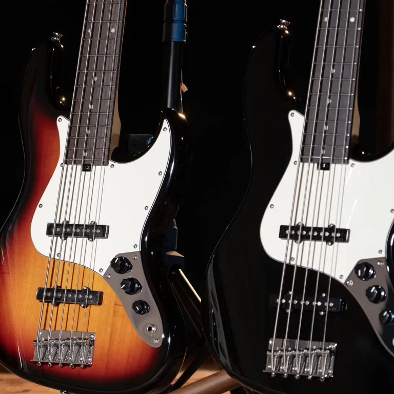 Two electric bass guitars with white pickguards resting side by side, one sunburst and one solid black, on stands in a dim setting. ขายราคาพิเศษ