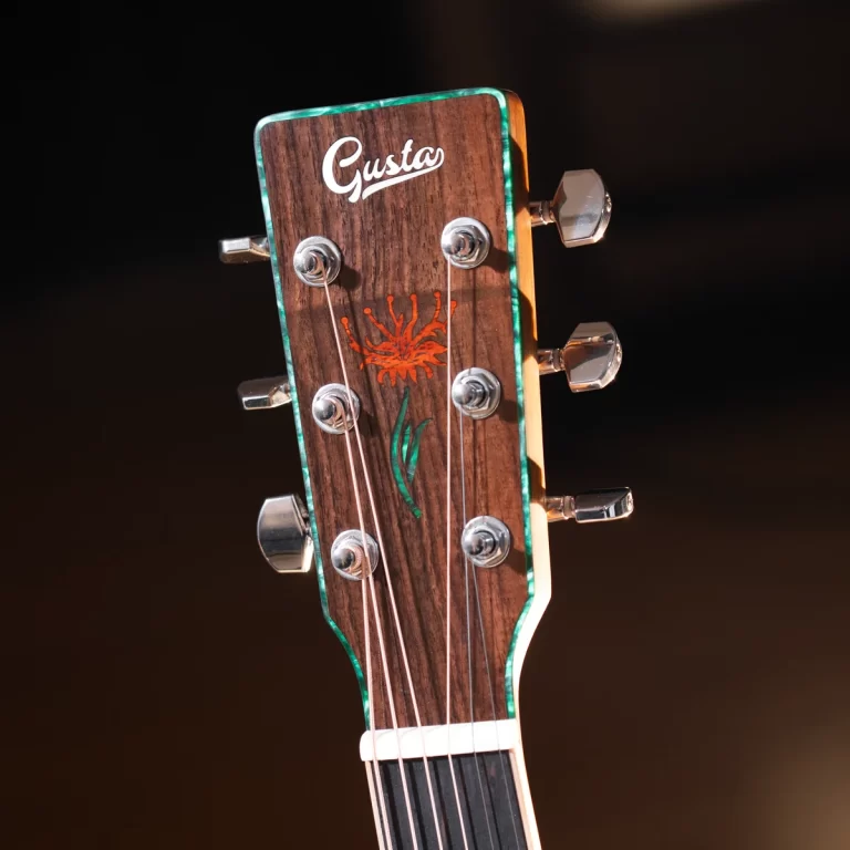 Close-up of a guitar headstock showing a wooden finish, silver tuning pegs, and a white 'Gusta' logo with a red flower design on the front panel ขายราคาพิเศษ