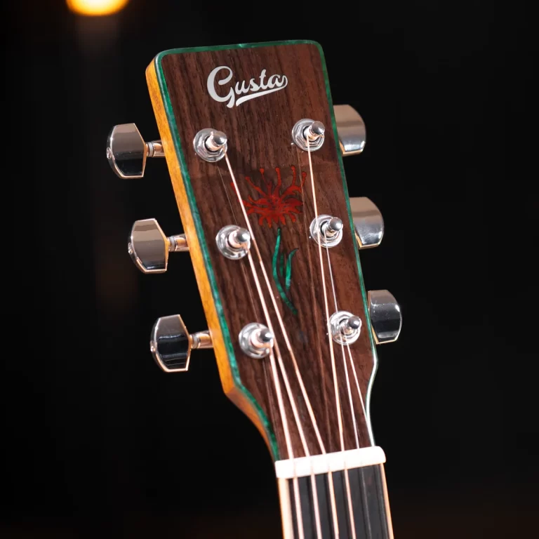 Close-up of a wooden guitar headstock with six chrome tuning pegs, strings, and a decorative inlay reading 'Gusta' against a dark background. ขายราคาพิเศษ