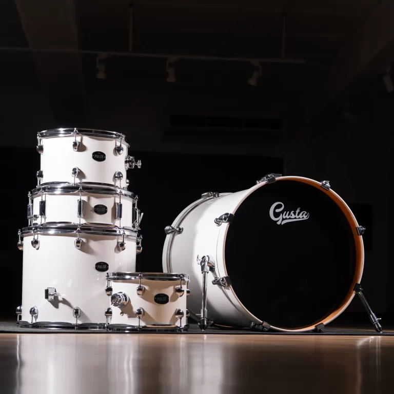 White drum kit on a dark stage, bass drum with 'Gusta' logo and chrome hardware on a wooden floor. ขายราคาพิเศษ