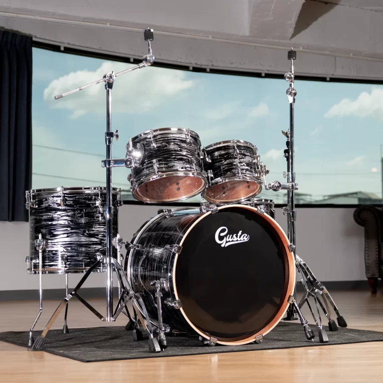 Full drum kit on a wooden floor with a black bass drum labeled 'Gusta' and chrome hardware in a rehearsal space. ขายราคาพิเศษ