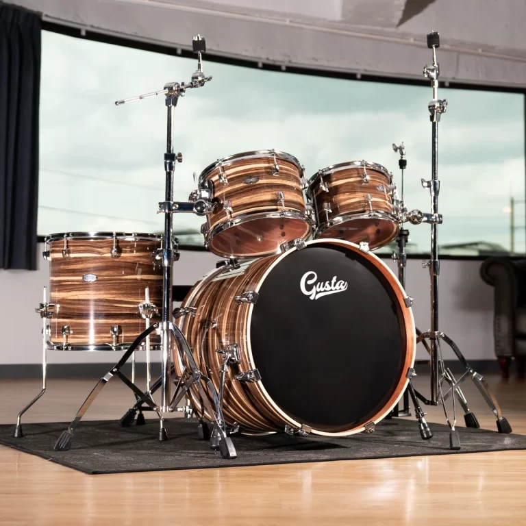 Five-piece wooden drum kit with chrome hardware on a black rug, bass drum bearing the Gusta logo. ขายราคาพิเศษ