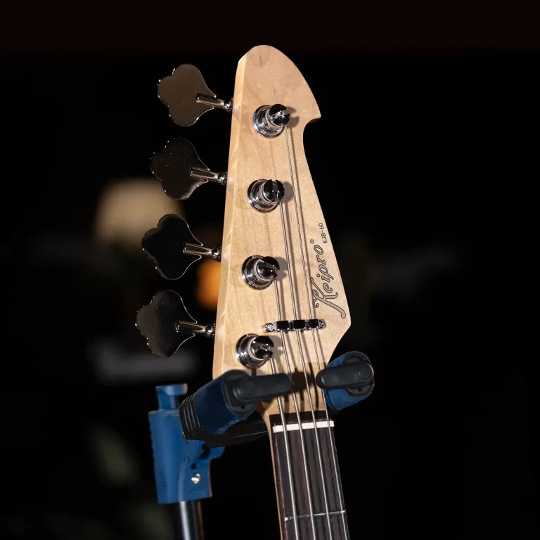 Close-up of a light-colored bass guitar headstock with four tuning pegs and strings, mounted on a stand against a dark background. ขายราคาพิเศษ
