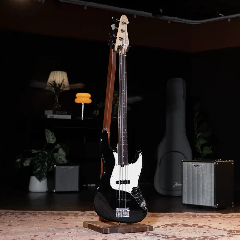 Black electric bass guitar on a wooden stand, centered on a rug, with an amplifier and guitar case in a dim studio setting. ขายราคาพิเศษ