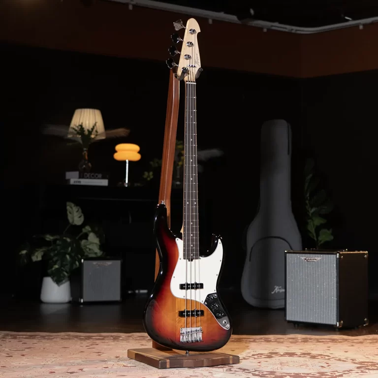 Sunburst electric bass guitar on a wooden stand in a dim room with amplifiers and a guitar case in the background ขายราคาพิเศษ