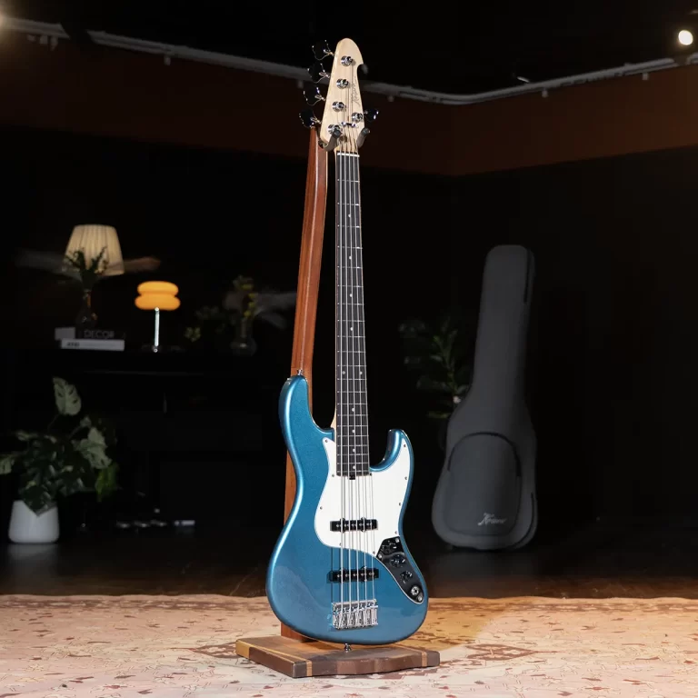 Blue electric bass guitar on a wooden stand in a dim studio, featuring a white pickguard and chrome bridge with a case in the background. ขายราคาพิเศษ