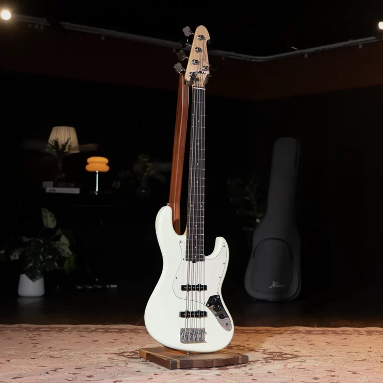 Cream-colored four-string electric bass guitar on a wooden stand in a dim room with a case in the background ขายราคาพิเศษ