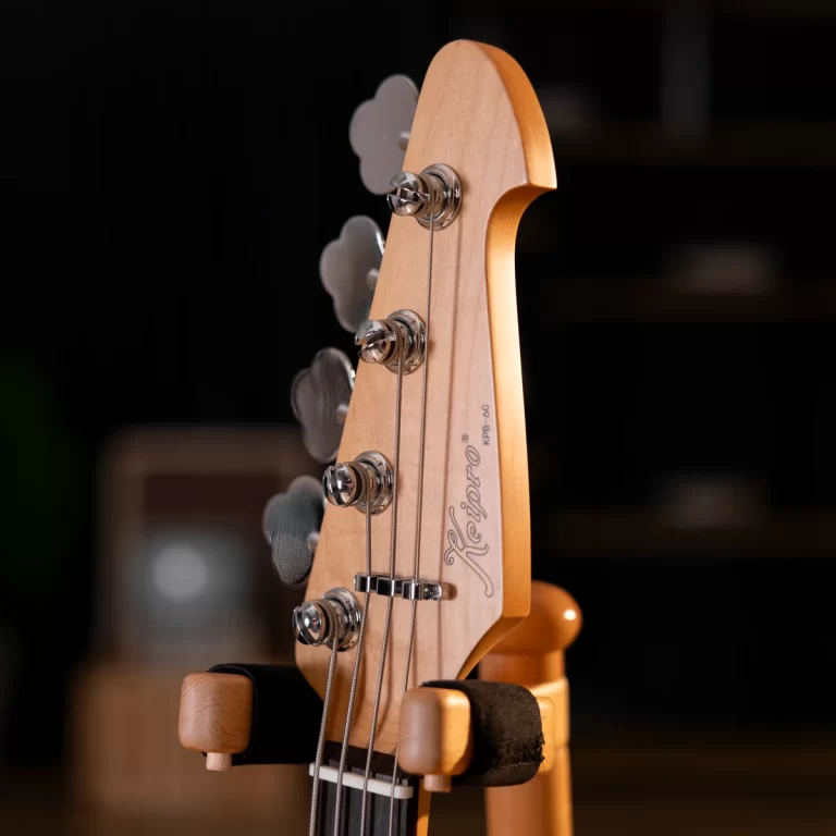 Close-up of a Fender bass guitar headstock with tuning pegs and strings, mounted on a wooden stand against a dark background ขายราคาพิเศษ