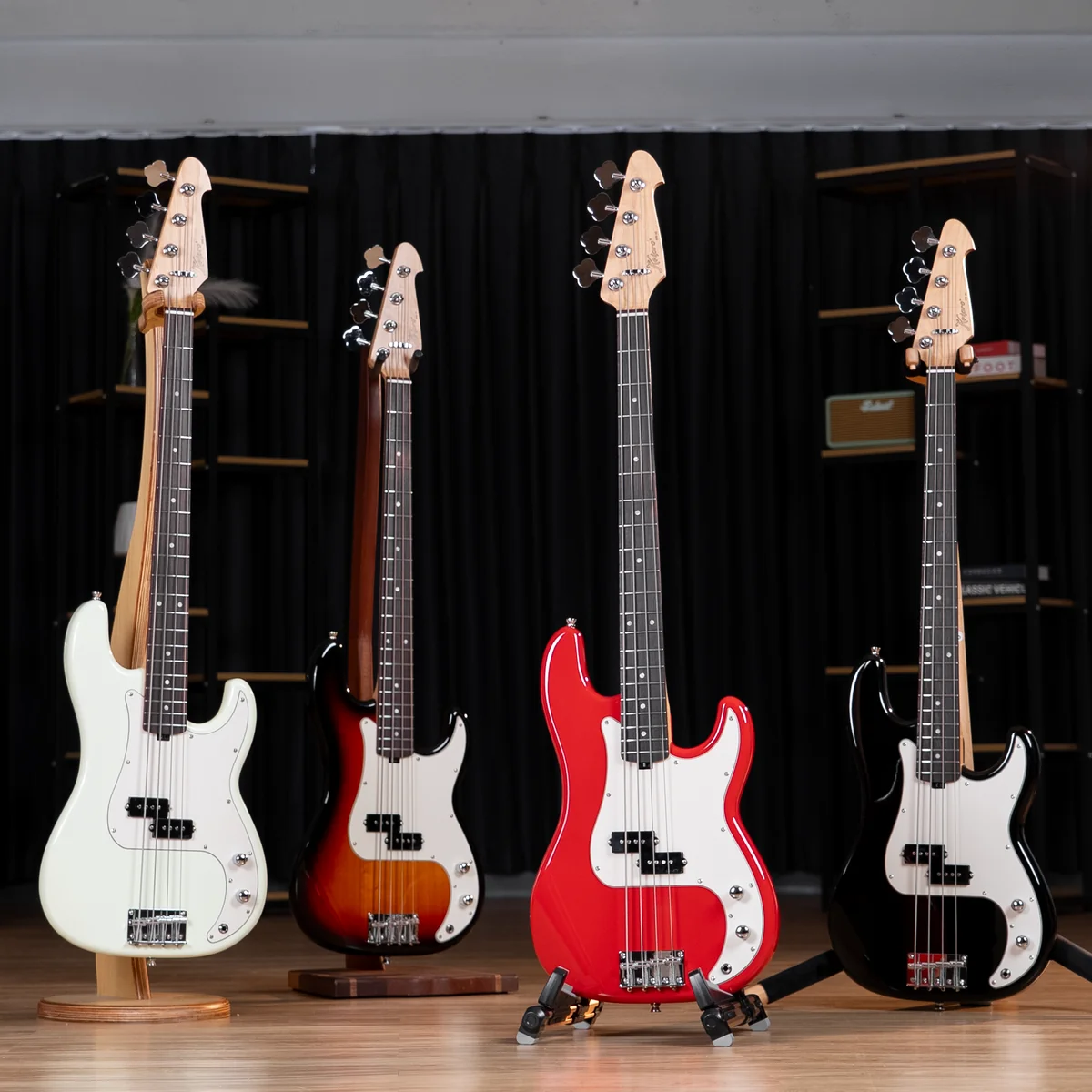 Four electric bass guitars on stands lined up in a music shop, white, sunburst, red, and black models against a black curtain backdrop on a wooden floor, showcasing diverse finishes.