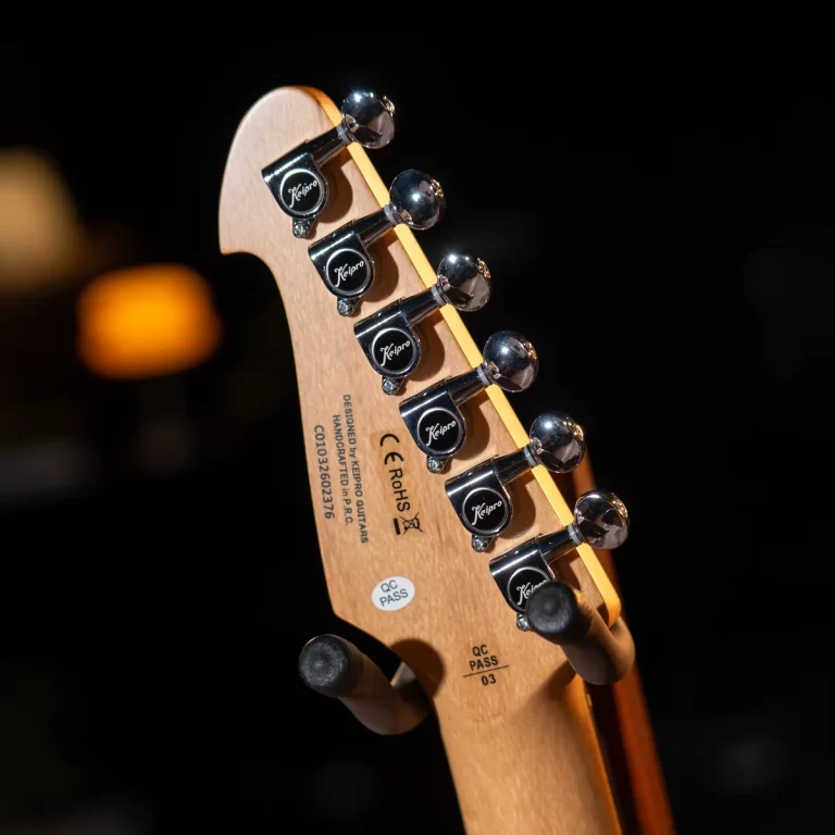 Close-up of a guitar headstock with six chrome tuning pegs, brand 'Keipro' visible on each tuner, warm background lights blurred. ขายราคาพิเศษ