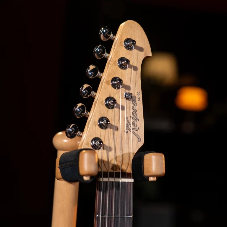 Close-up of a wooden guitar headstock with chrome tuning pegs on a stand, against a dark background. ขายราคาพิเศษ