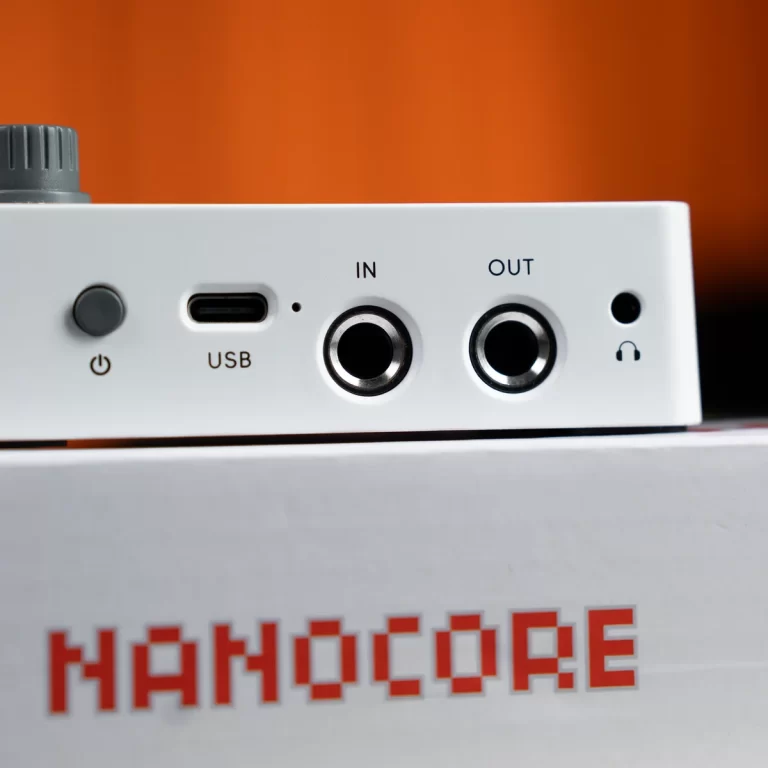 Close-up of a white device front panel with USB-C port, IN and OUT jacks, a power button, and a headphone jack against an orange background; NANOCORE text on the box below. ขายราคาพิเศษ
