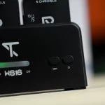Close-up of a black charging dock with two rounded buttons and a power icon; a white chain-link symbol suggesting pairing or connectivity, with a blurred device in the background. ขายราคาพิเศษ