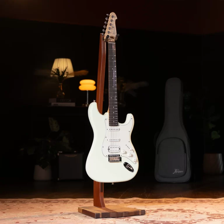 White Fender-style electric guitar on a wooden stand in a dim room with a guitar case in the background. ขายราคาพิเศษ