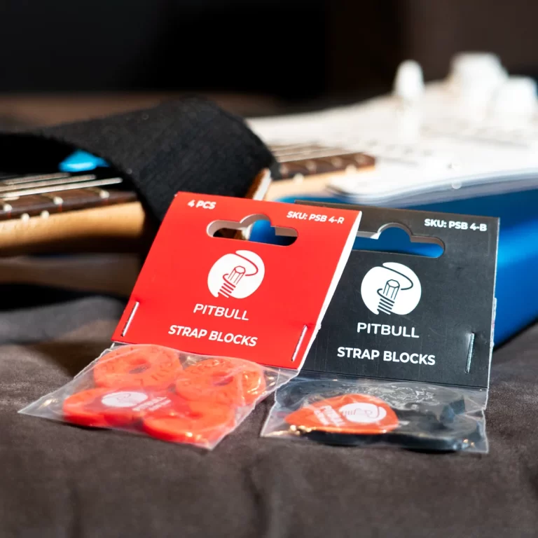 Two packages of Pitbull strap blocks (red and black) displayed on a table in front of a guitar case or neck. ขายราคาพิเศษ