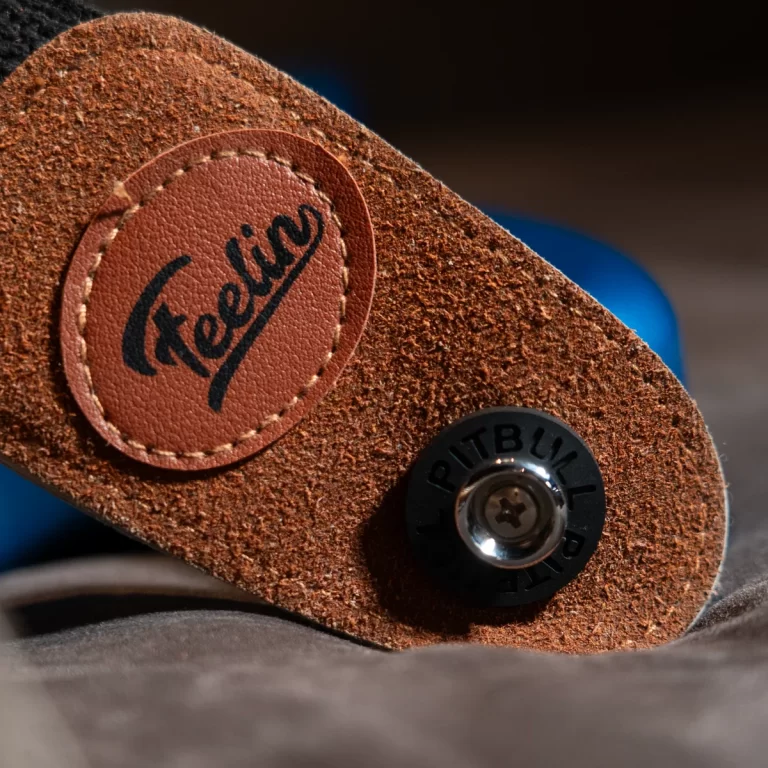 Close-up of a brown suede strap with a stitched leather patch reading 'Felina' and a small black circular fastener beside it on a textured surface. ขายราคาพิเศษ