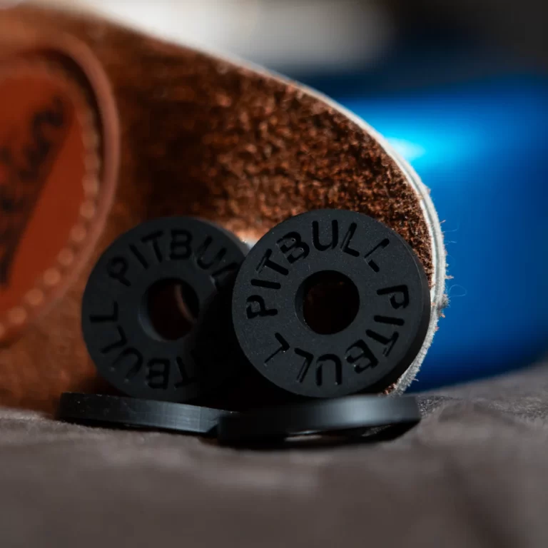Close-up of black rubber circular pieces with 'PITBULL' embossed around the edge, resting on a brown leather surface. ขายราคาพิเศษ