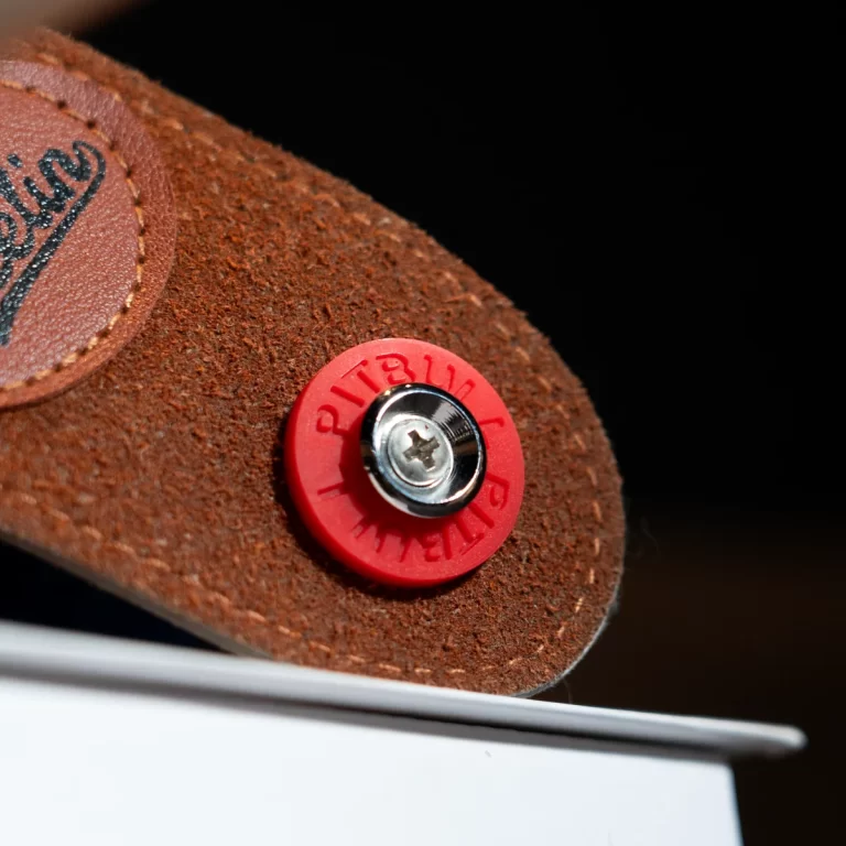 Close-up of a brown suede shoe with a red circular rivet and metal screw, set against a dark background and a white edge nearby. ขายราคาพิเศษ