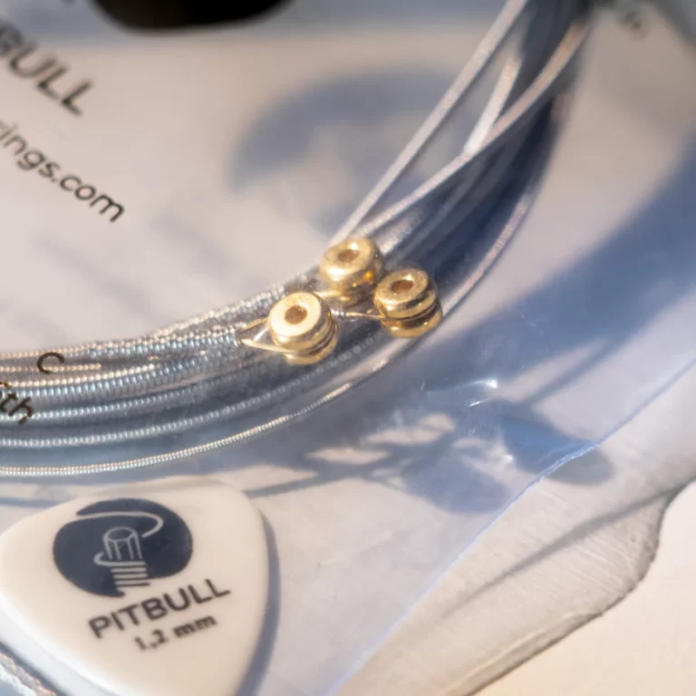 Close-up of silver guitar strings wrapped around a curved metal edge with a Pitbull 1.3 mm guitar pick in the foreground on a shiny surface. ขายราคาพิเศษ