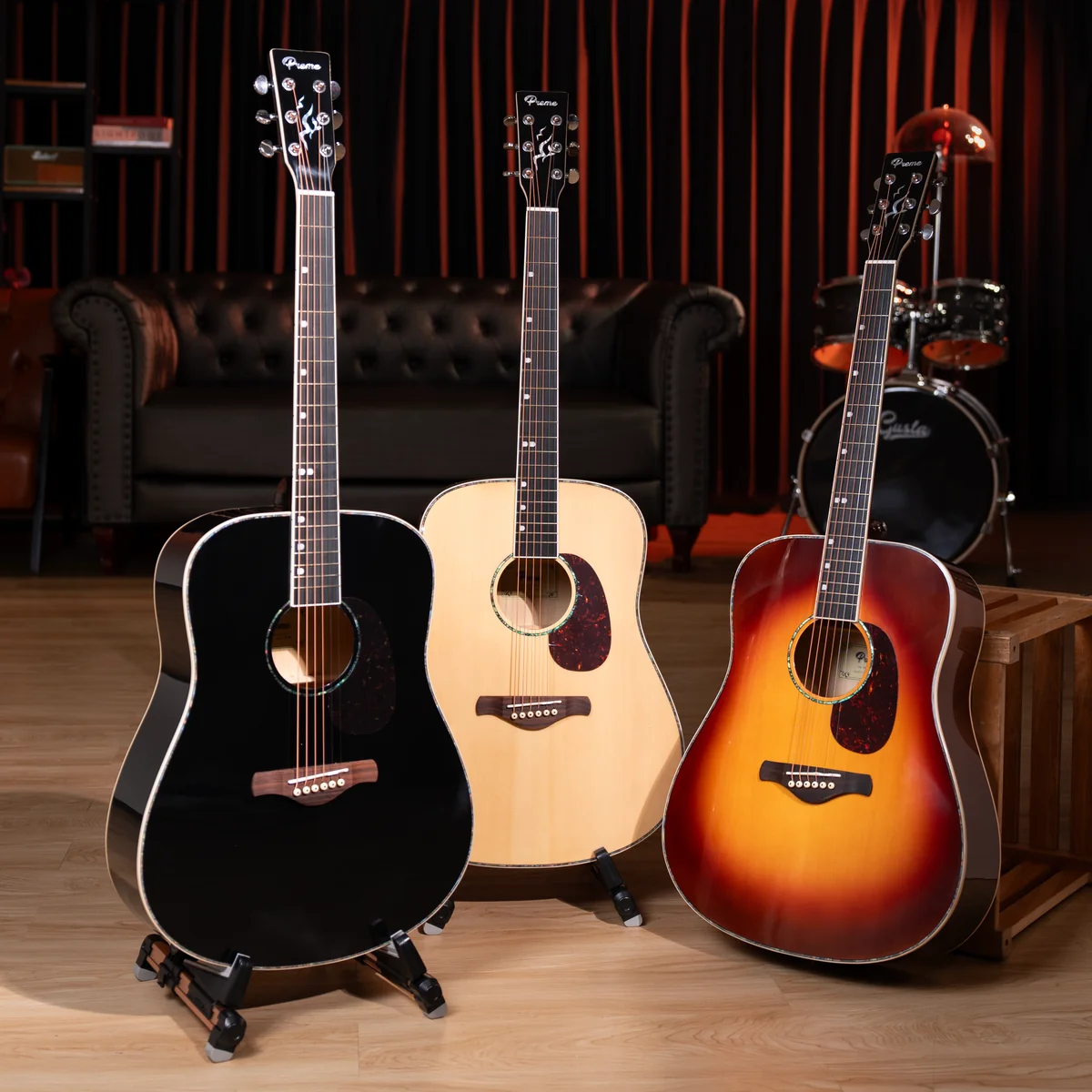Three acoustic guitars on stands lined up on a wooden floor in a cozy studio with a black, natural, and sunburst finish set against a dark sofa and drum kit background.