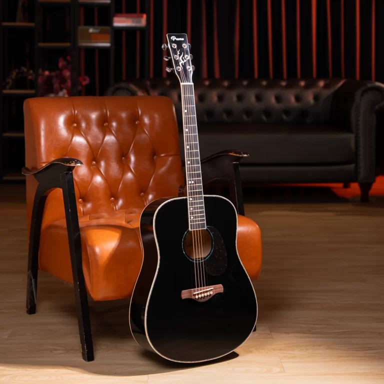 Black acoustic guitar resting against an orange leather armchair in a dimly lit studio/lounge setting. ขายราคาพิเศษ