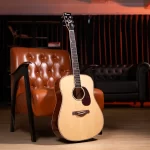 Acoustic guitar resting on the floor beside a tufted brown leather chair in a cozy studio setting ขายราคาพิเศษ