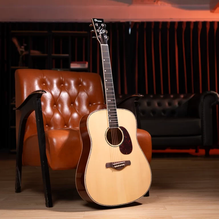 Acoustic guitar resting on the floor beside a tufted brown leather chair in a cozy studio setting ขายราคาพิเศษ