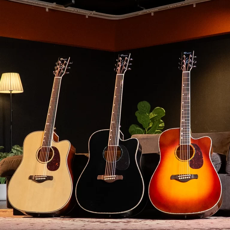 Three acoustic guitars stand side by side in a dim room: natural, black, and sunburst finishes from left to right, with a lamp and plant in the background. ขายราคาพิเศษ