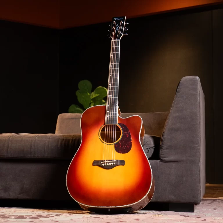 Sunburst acoustic guitar resting upright against a gray sofa in a dim living room setting. ขายราคาพิเศษ
